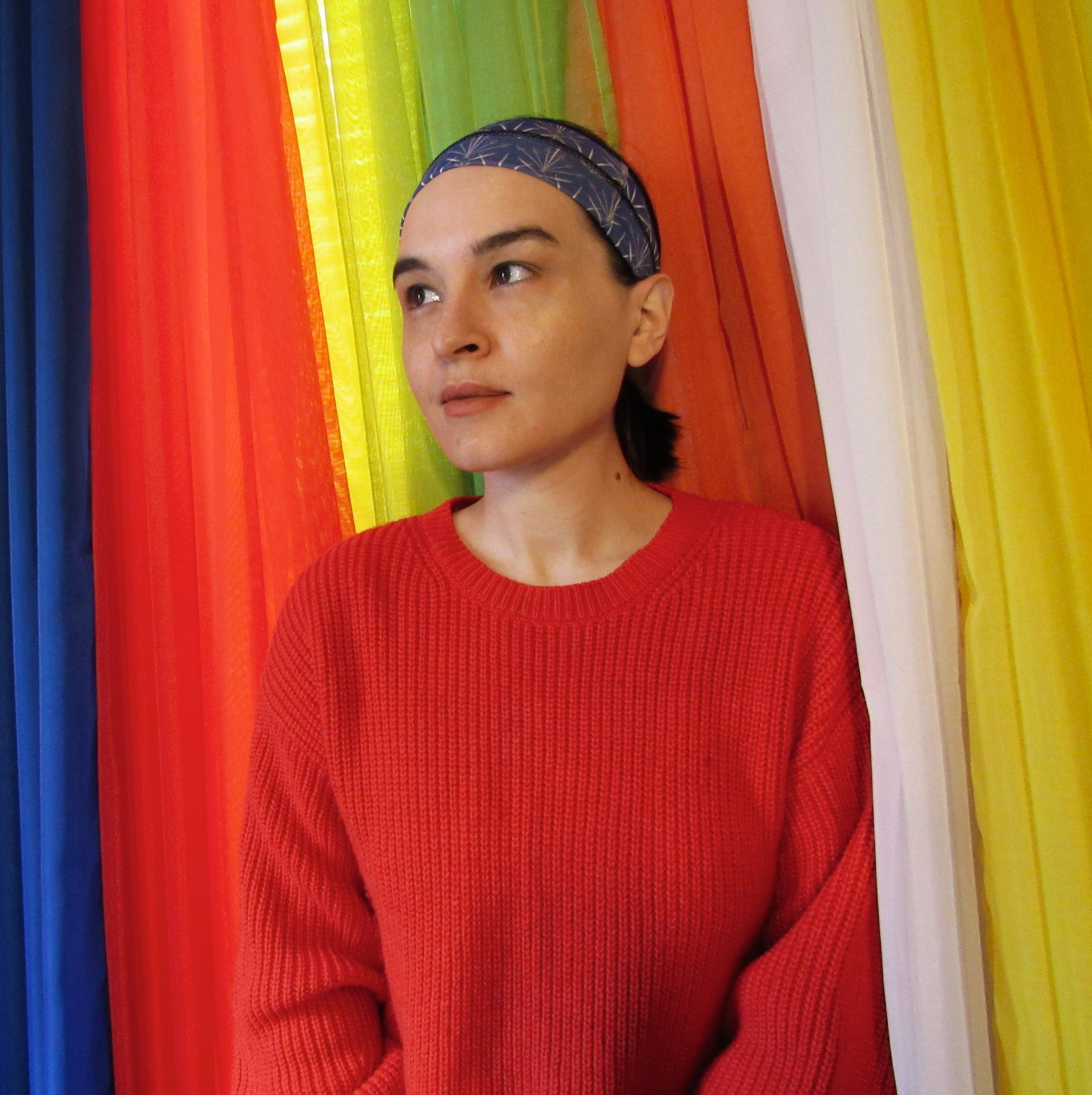 Portrait of Darcie Little Badger wearing a red sweater, standing against brightly-colored curtains