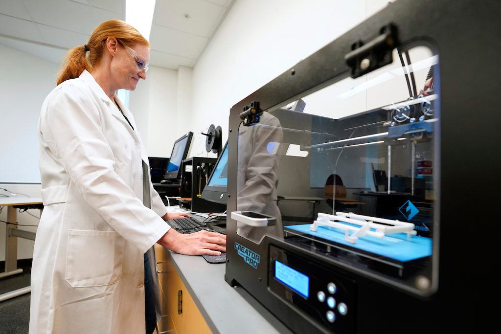 a photo of a smiling woman in a lab coat and safety glasses operating a computer next to a large 3d printer