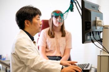 a photo of a man in a lab coat staring at a computer screen while a young woman in the background sits with a series of cables attached to her head