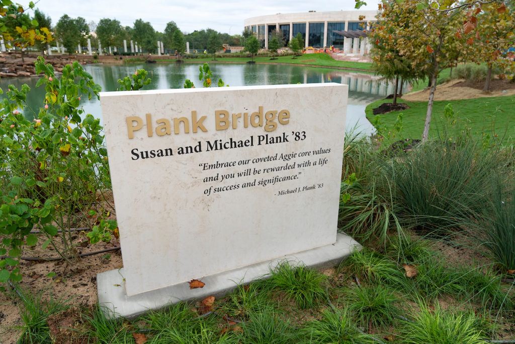 A marker that reads "Plank Bridge" with a pond and the Association of Former Students building in the background