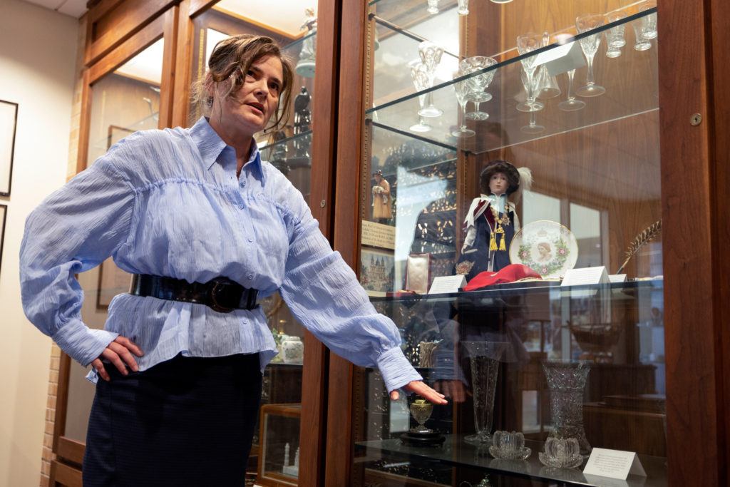 a photo of a woman in a blue shirt gestures towards a glass case containing various pins, medals, glassware and other items