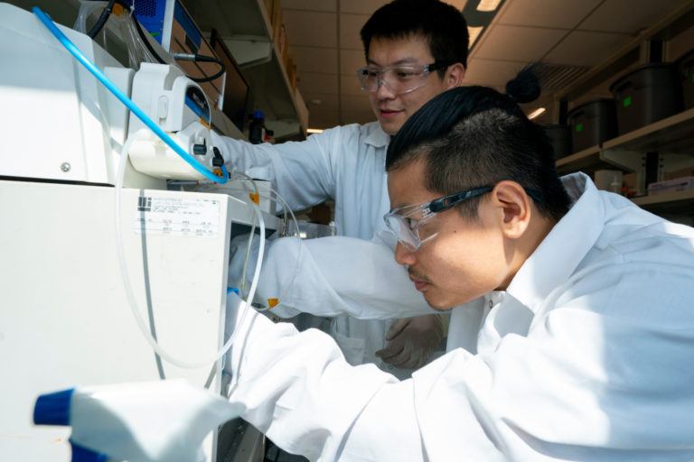 a photo of two men in lab coats and safety glasses looking at lab equipment