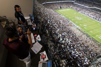 in a booth overlooking the crowd and football field at kyle field, two men and a woman stand in a broadcasting booth