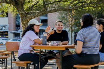 three female students and one male student sit at a picnic table under an oak tree wrapped in holiday lights