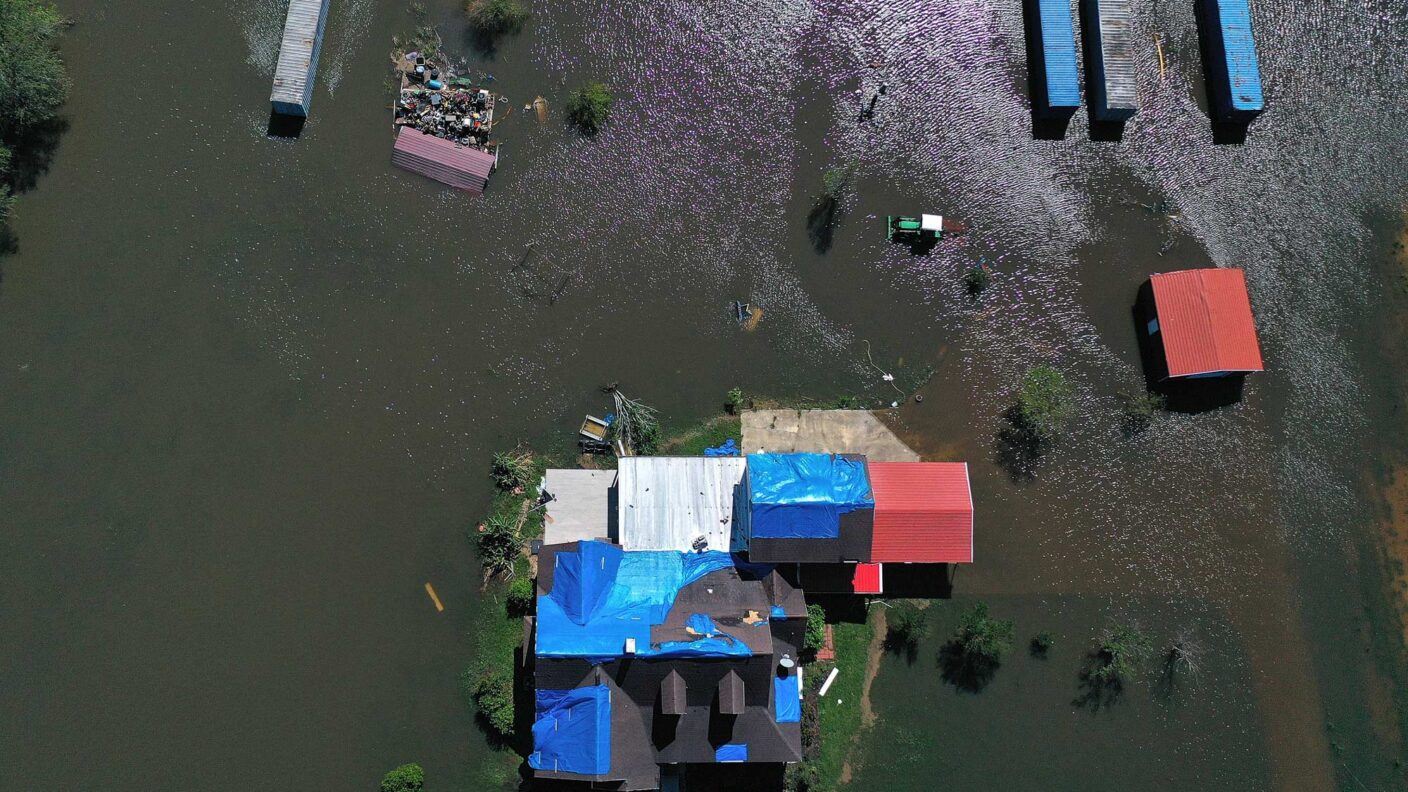 a top-down photo of a house sitting just above surrounding flood waters. The roof of the house has been patched up with blue tarps. Behind the house, a tractor, outbuilding and several storage containers sit in the floodwater.