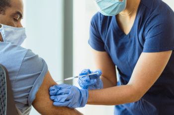 An unrecognizable adult female nurse gives her mature adult male patient a flu shot in his arm. He watches intently. Both the nurse and the patient wear protective masks.