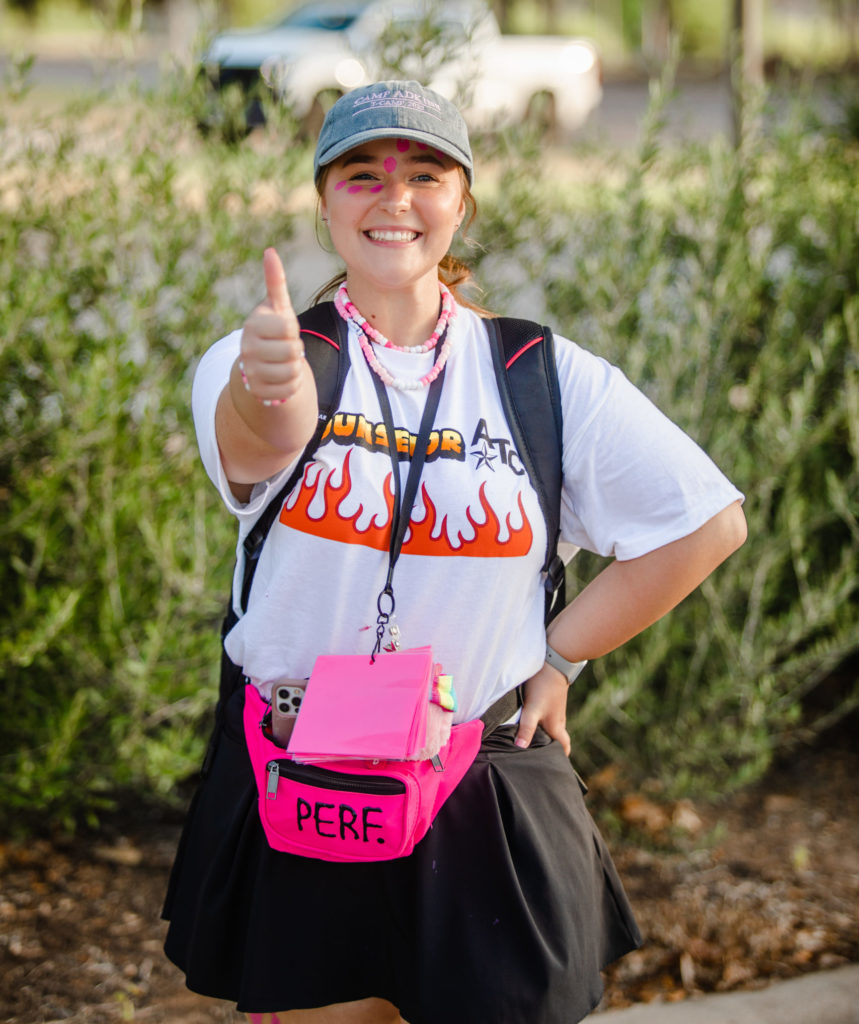 a photo of a young woman in a black skirt and white shirt smiling and giving the thumbs up. she has dots of pink paint on her face and is wearing a pink fanny pack and necklaces.