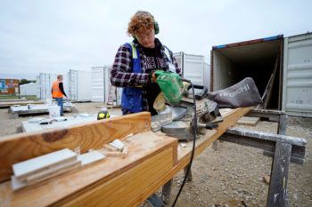 a photo of a young man in a flannel shirt and safety gear cutting wood with a large saw