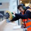 a photo of a young man in eyeglasses and safety gear operating a large saw. bits of sawdust fly in the air around him.