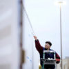 a photo of a young man in a dark blue and maroon jacket standing on a ladder applying white paint to the side of a shipping container