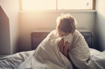 A sick boy sitting in a bed covered in blankets while blowing his nose