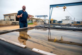 A man standing next to a large vat of salvaged wood that can be seen submerged under water