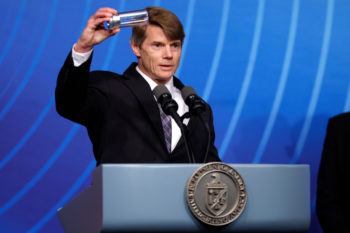 Marvin Adams standing behind a podium holding up a cylinder in one hand