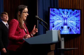 a photo of a woman in a reddish jacket speaking at a podium in front of a screen displaying a series of tubes and bright light. Behind her in the frame, a man in a suit watches.