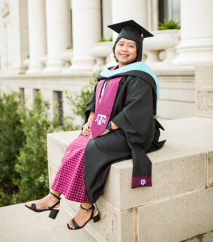 a photo of a woman in a dress, heels, and a graduation cap and gown sitting outside the administration building on the Texas A&M campus