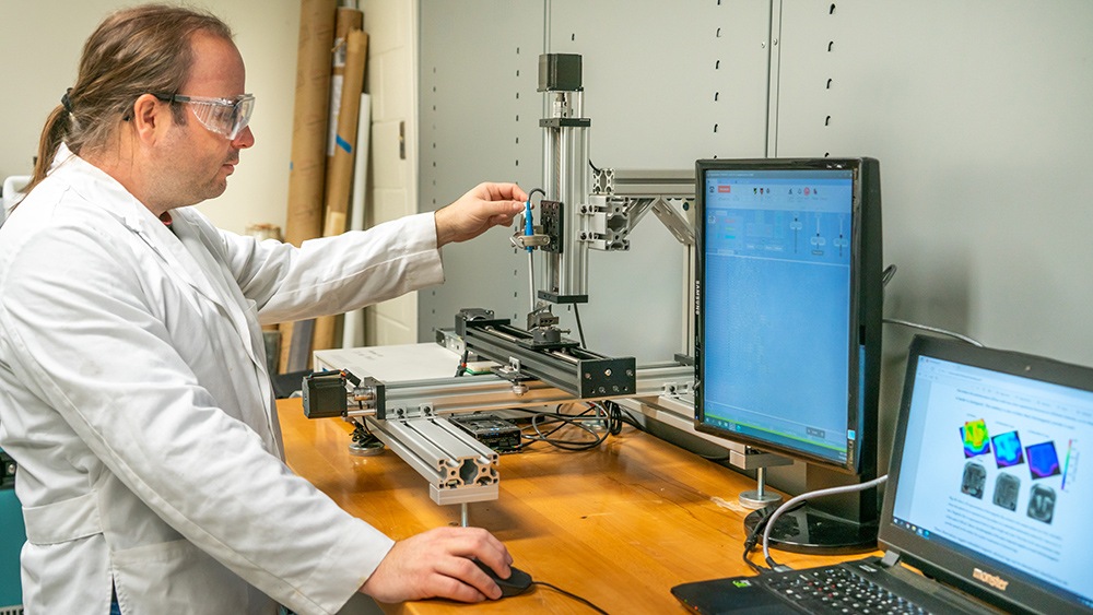 a photo of a man in a lab coat and safety goggles operating a machine next to a couple of computer screens