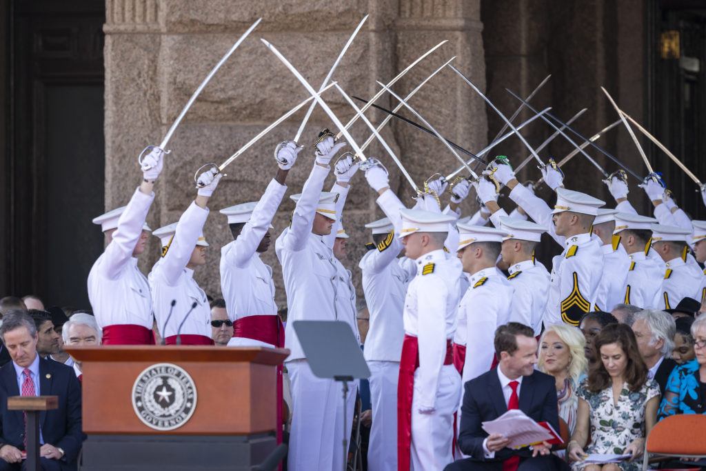 a photo of cadets in white uniforms holding up sabers to form an archway, creating a path to a podium in front of the state capitol building