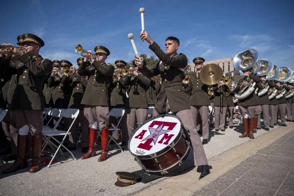 a photo of uniformed cadets playing instruments at the state capitol. in the center of the photo is a bass drummer with his hat off and his arms raised