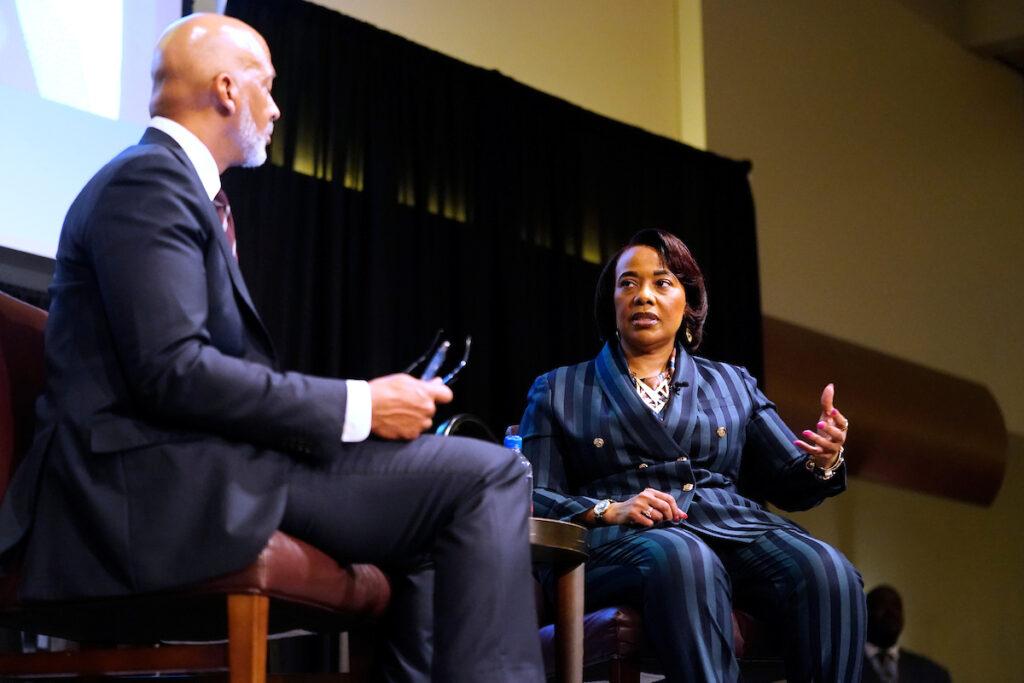 a photo of a man and a woman in suits sitting next to each other on a stage. the woman is looking at the man and speaking while holding out her hand.