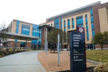 A view from outside ILSQ’s south entrance, showcasing the building’s front lawn and outdoor study space, with the building itself standing in the background. A sign on the right side of the frame displays the name of the building.