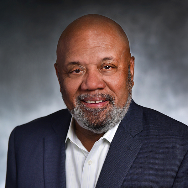 a photo of a man with a black and grey beard smiling into the camera. he is wearing a navy blue sports coat over a white button-up shirt.