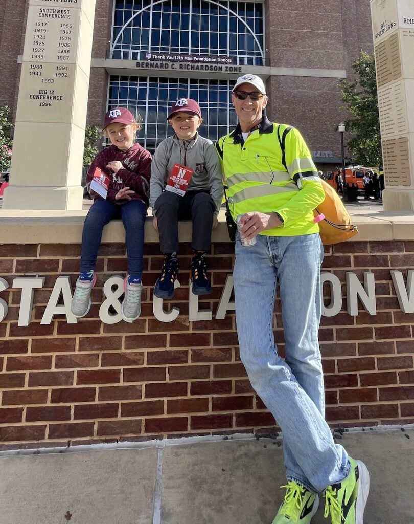 a photo of a man in a bright yellow safety shirt next to two small children in Texas A&M caps in front of Kyle Field