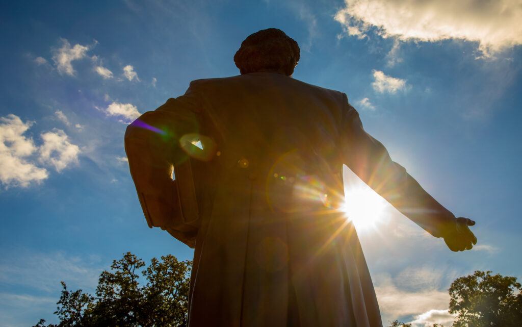 a photo of the statue of Matthew Gaines from behind with the sun in front, shooting beams of light under his outstretched arm