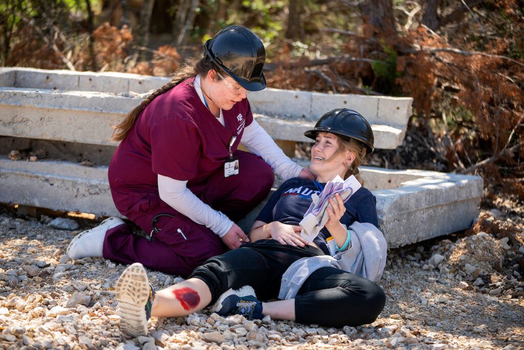 a photo of a student in maroon scrubs kneeling down and speaking with a woman laying on the ground with red makeup on her arms and legs