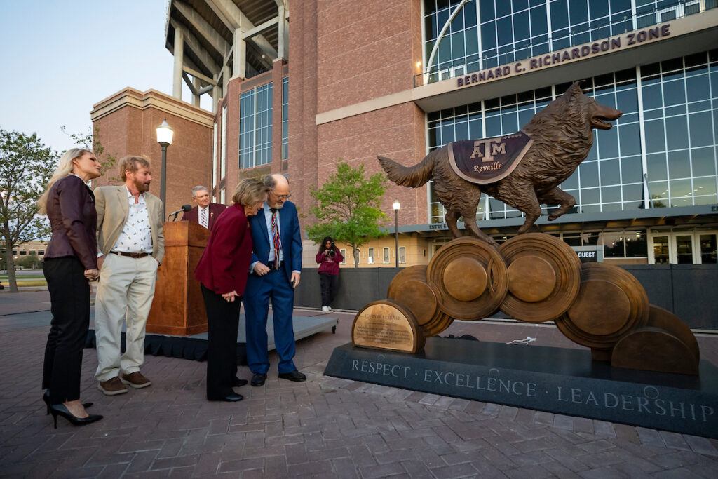 People stand to the side of a large bronze statue of Reveille in front of Kyle Field