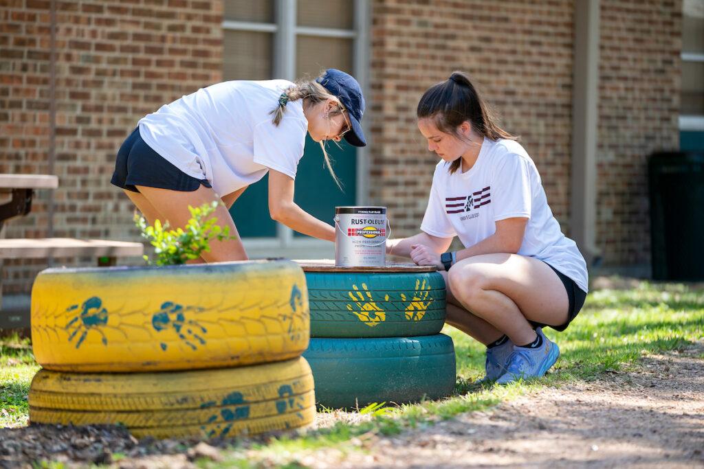Two female students paint tires that have been converted into planters outside an elementary school