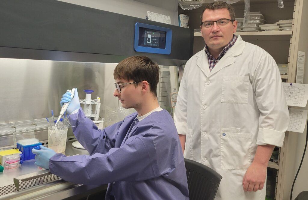a photo of a man in a lab coat standing next to a younger man in a blue protective gown working under a fume hood