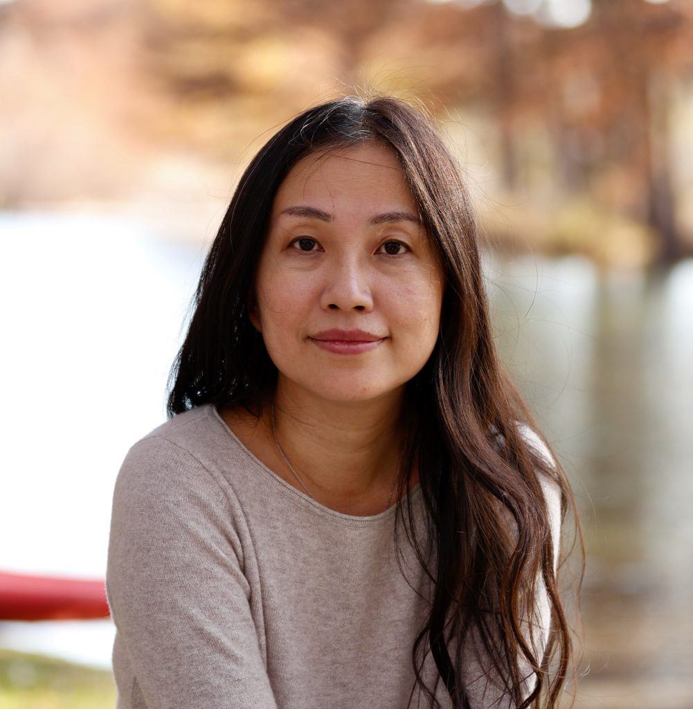 a headshot of a woman with long dark hair wearing a sweater with water and trees in the background