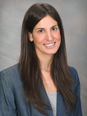 a headshot of a woman with long dark hair wearing a blazer against a grey backdrop