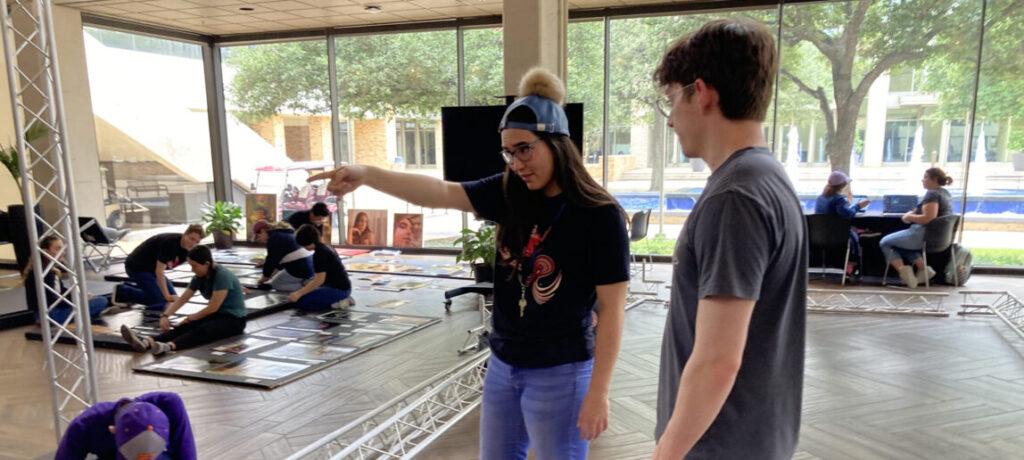 a photo of a young man and a young woman overseeing a group of students placing paintings and other works of art on boards for display