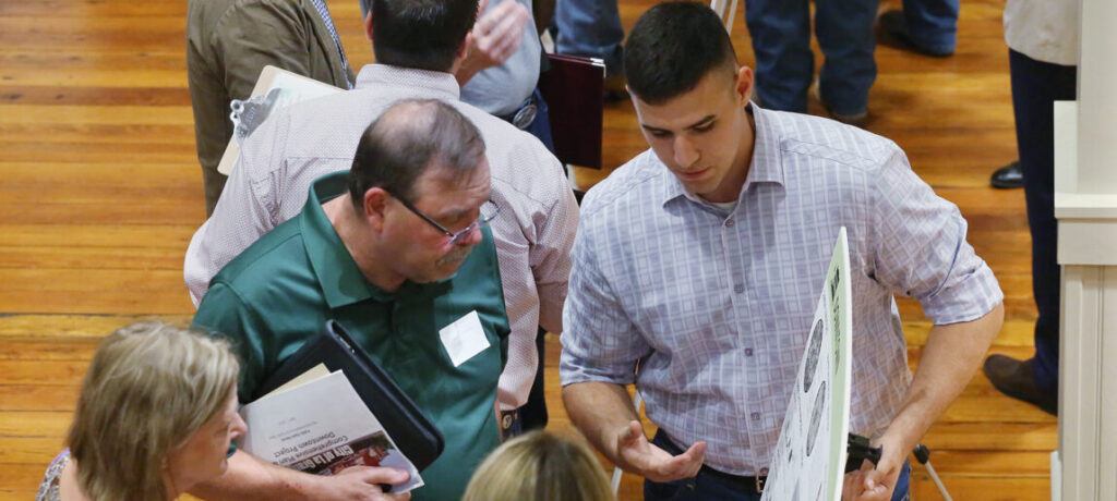 a photo of a young man in a checkered shirt showing a poster on an easel to a group of older people. the contents of the poster are not readable from this angle.