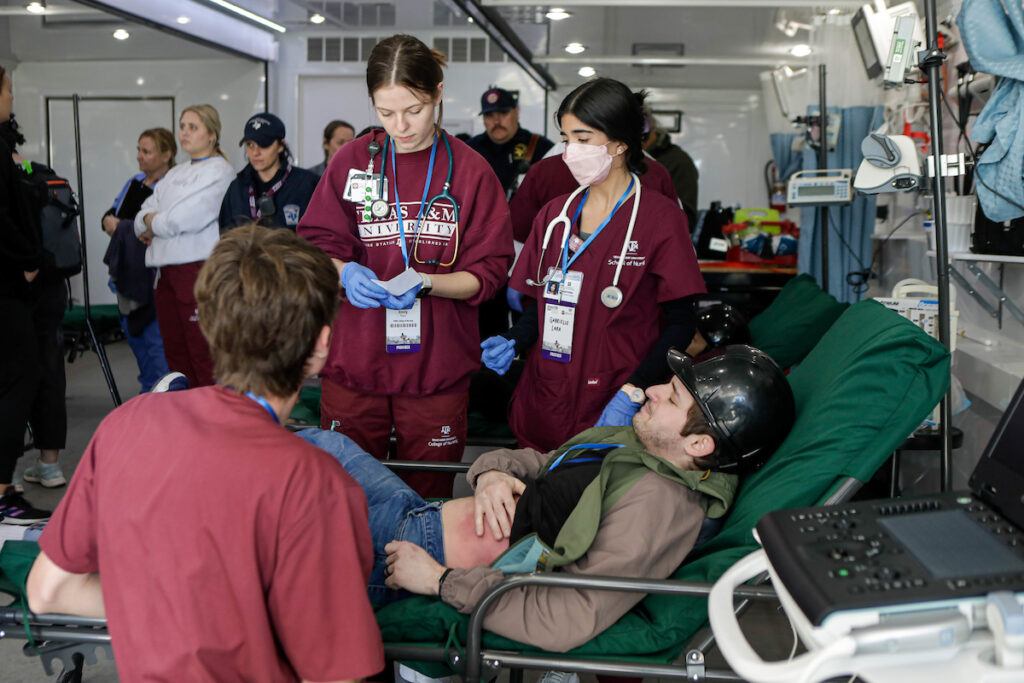 a photo of three women in maroon scrubs around a man laying down. he clutches a fake bruise on his abdomen.