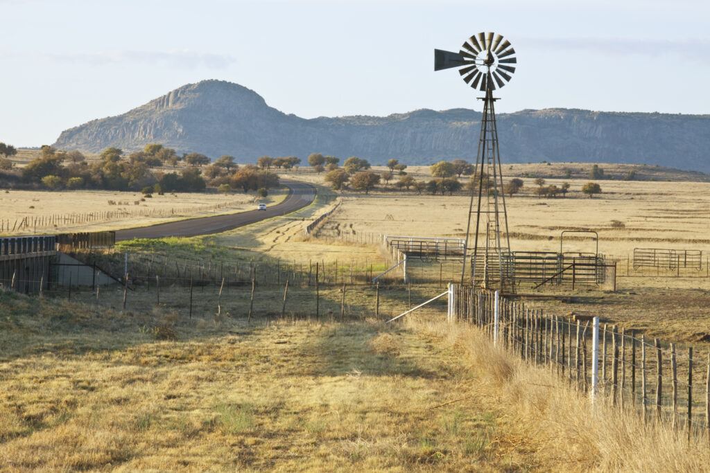 Fence-line leading to West Texas Ranch corrals and windmill, with highway running by. In early morning light.