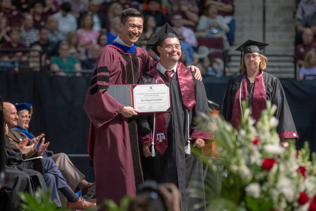 A man in a maroon robe places his arm around a young man in a graduation gown and smiles, handing him a certificate