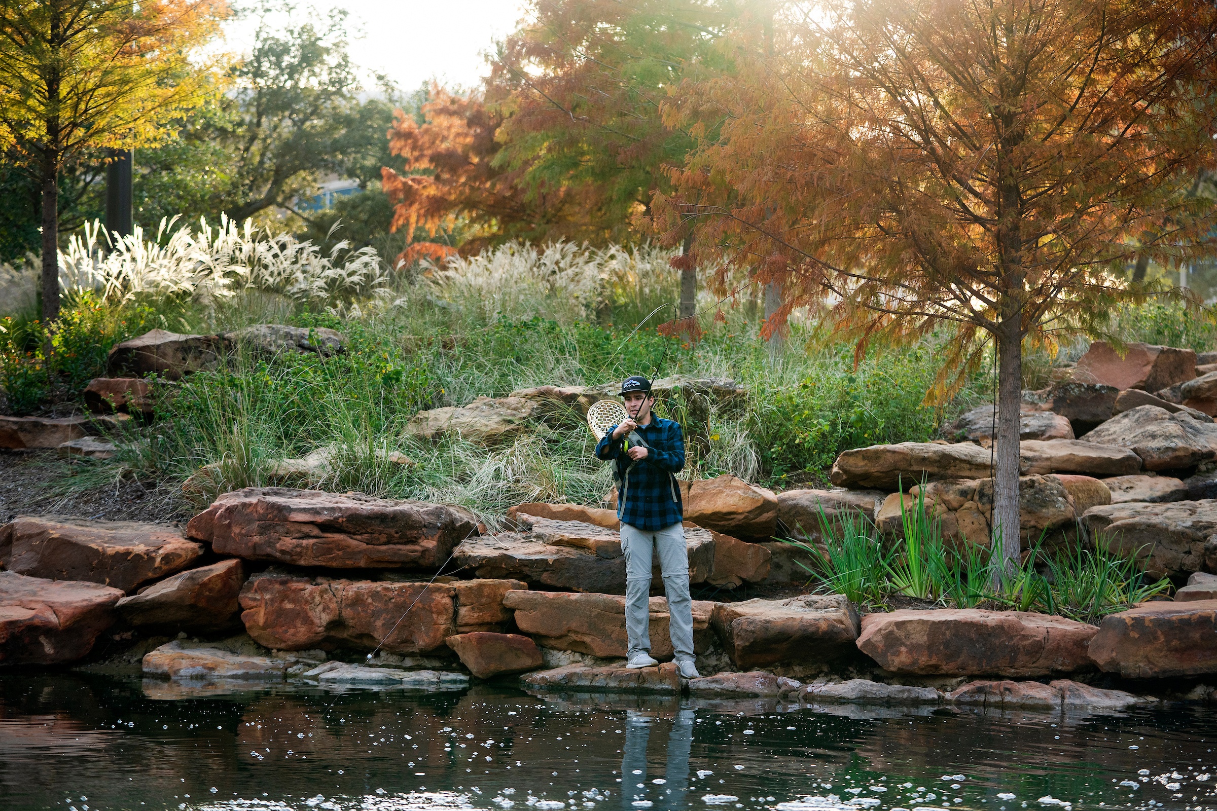 a photo of a young man in a flannel shirt fishing in a pond with the sun shining through cypress trees behind him