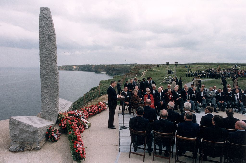 a photo of Ronald Regan delivering a speech on top of a high cliff next to a stone monument, as various government officials and other attendees watch