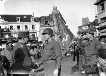 a black and white photo of a man in a french military uniform shaking hands with a man in an American military uniform in a town square as soldiers and civilians look on