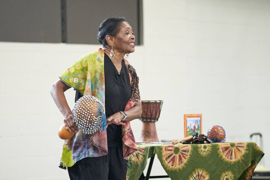 a photo of a woman holding a gourd instrument with a variety of instruments and other items behind her