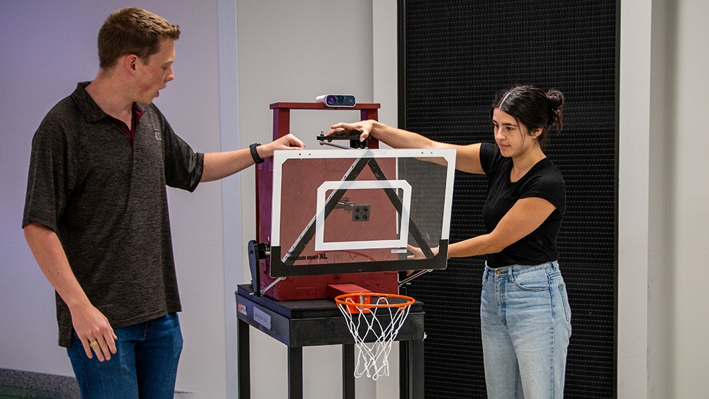 a young man and a young woman adjust a small basketball hoop sitting on a cart with a camera/sensor on top