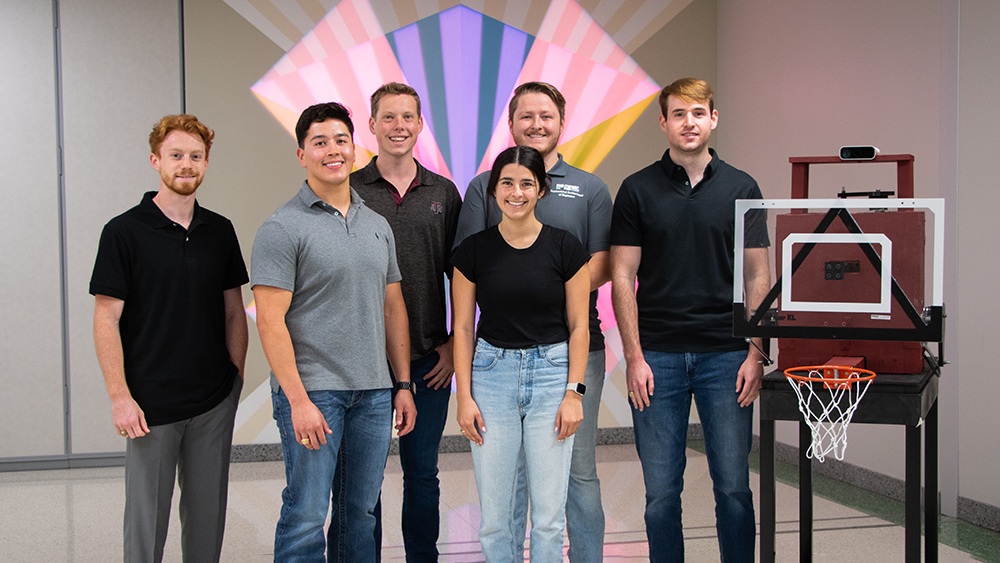 a photo of a group of students, five men and one woman, standing next to a small basketball hoop with a camera on top