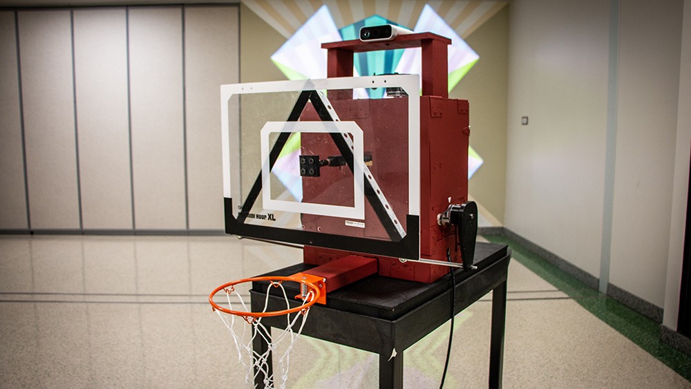 a small basketball hoop mounted on a maroon box with various wires and actuators coming out of it. A camera/sensor sits above the box.