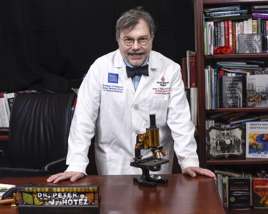 a portrait phot of a man with grey hair, glasses, a lab coat, and a bowtie standing at a desk with an old microscope in front of him