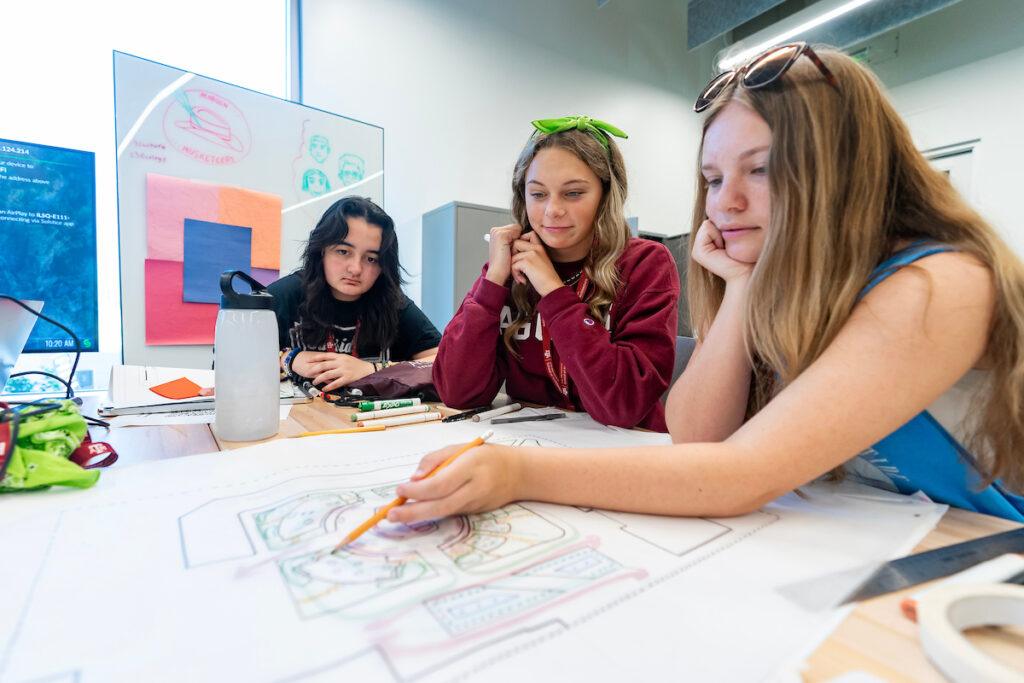 a photo of three young women looking at architectural plans on a table with a whiteboard and screen behind them