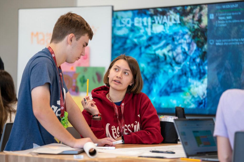 a young woman gestures with a pencil while speaking with a young man in a lab/classroom space