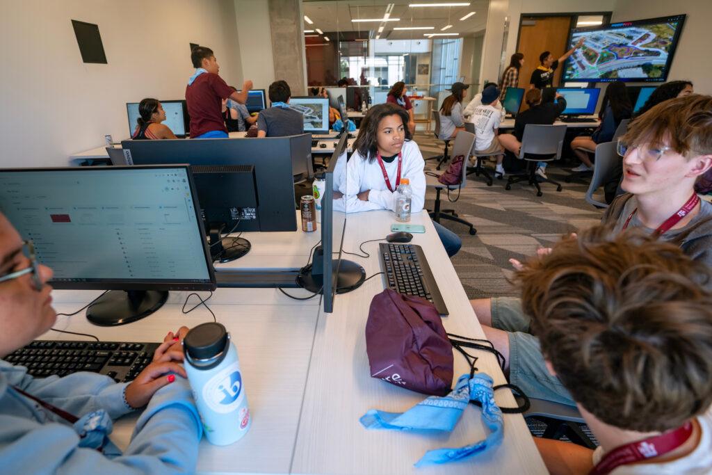 a photo of a young woman watching several other young people talk. all are seated around computers