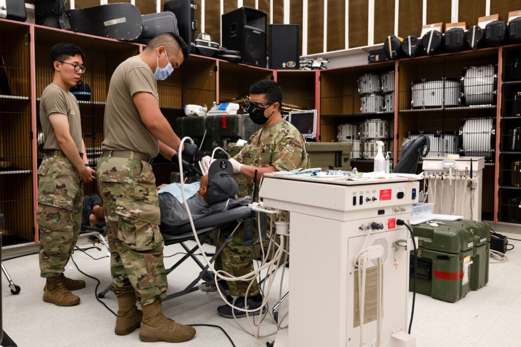 A photo of several people in army fatigues using various dental instruments a reclining man. In the background, drums and other instruments can be seen inside lockers.